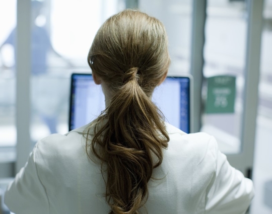 Woman doctor with long red hair using laptop in hospital, photographed from behind
