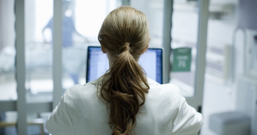 Woman doctor with long red hair using laptop in hospital, photographed from behind