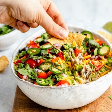 scooping healthy 7-layer dip out of a serving bowl with a tortilla chip