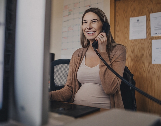 A woman who is pregnant talks on a landline phone in an office setting