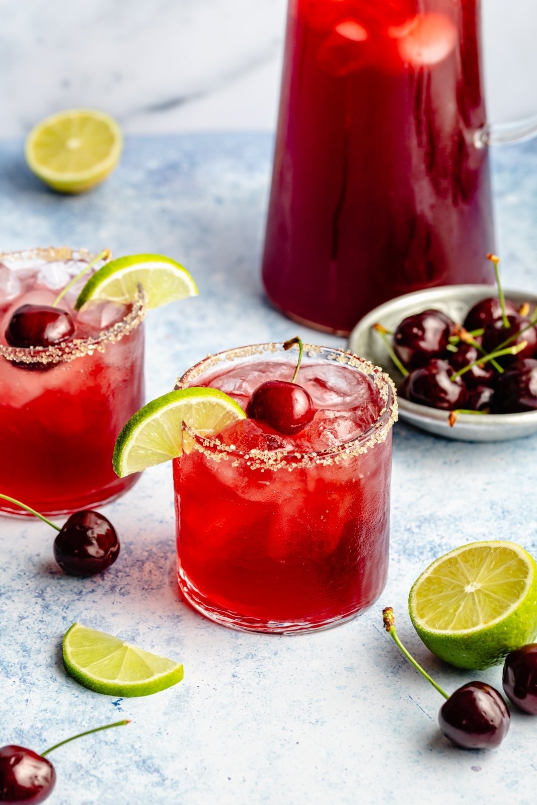 cherry lime cocktail in two glasses next to limes and cherries