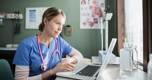 A woman wearing scrubs sits at a desk in a hospital setting and uses a laptop computer.