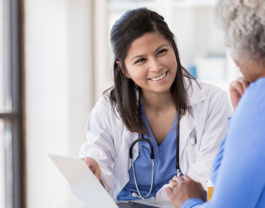 A young nurse uses laptop at a desk, while talking and smiling with a senior woman.