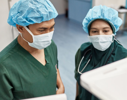 Elevated view of male surgeon and female anesthetist wearing green scrubs, caps, and masks checking computer monitors in operating room.